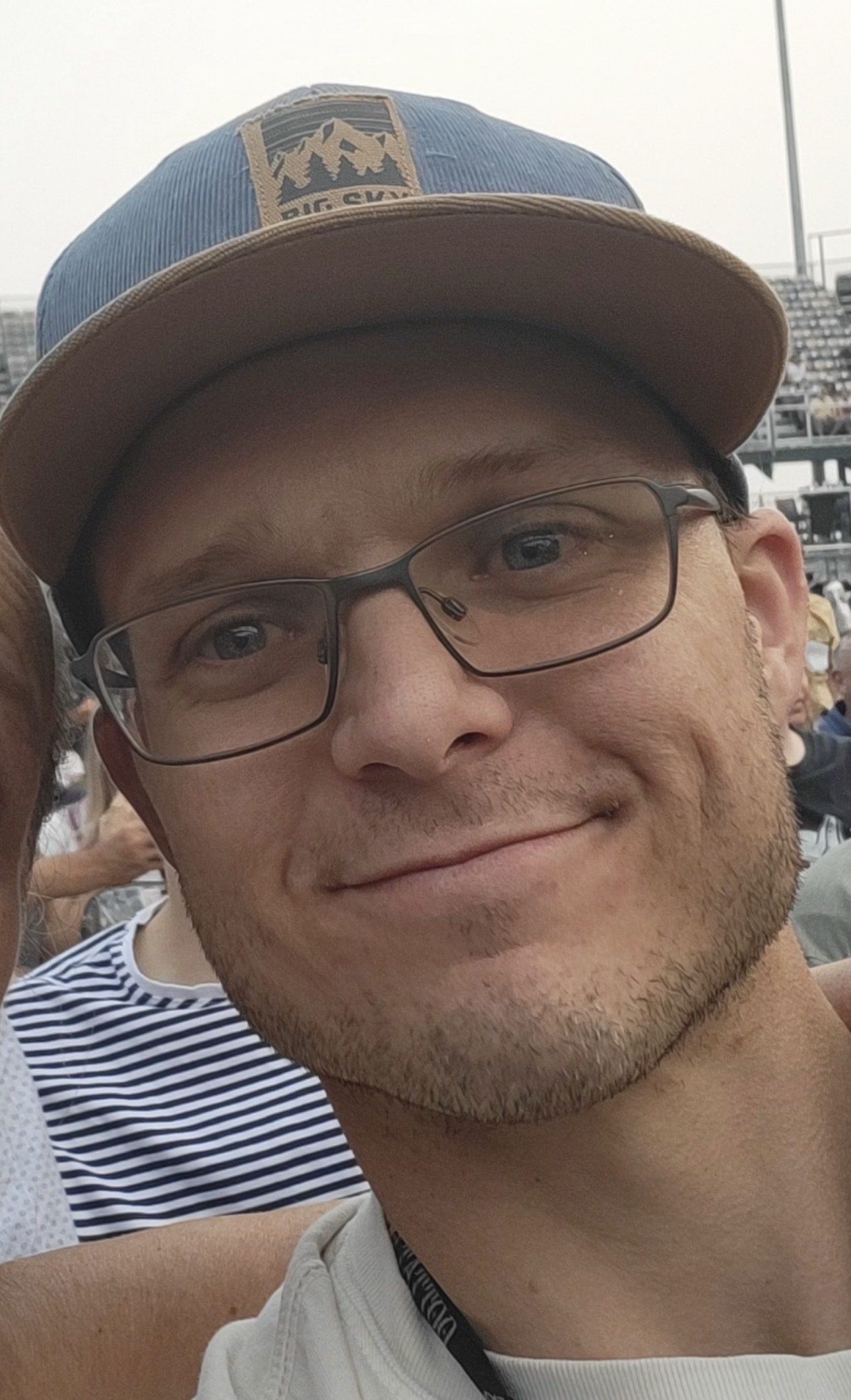 A man wearing glasses and a hat stands in a crowd at an outdoor sporting event.