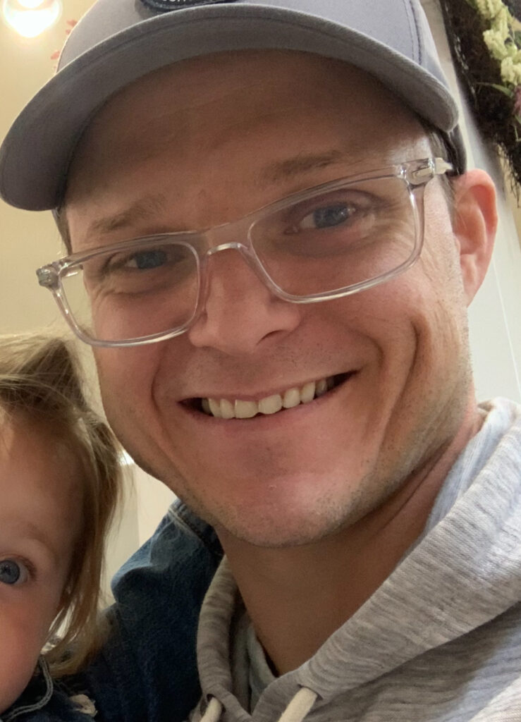 A man in glasses and a hat gently holds a little girl, both smiling in a warm and joyful moment together