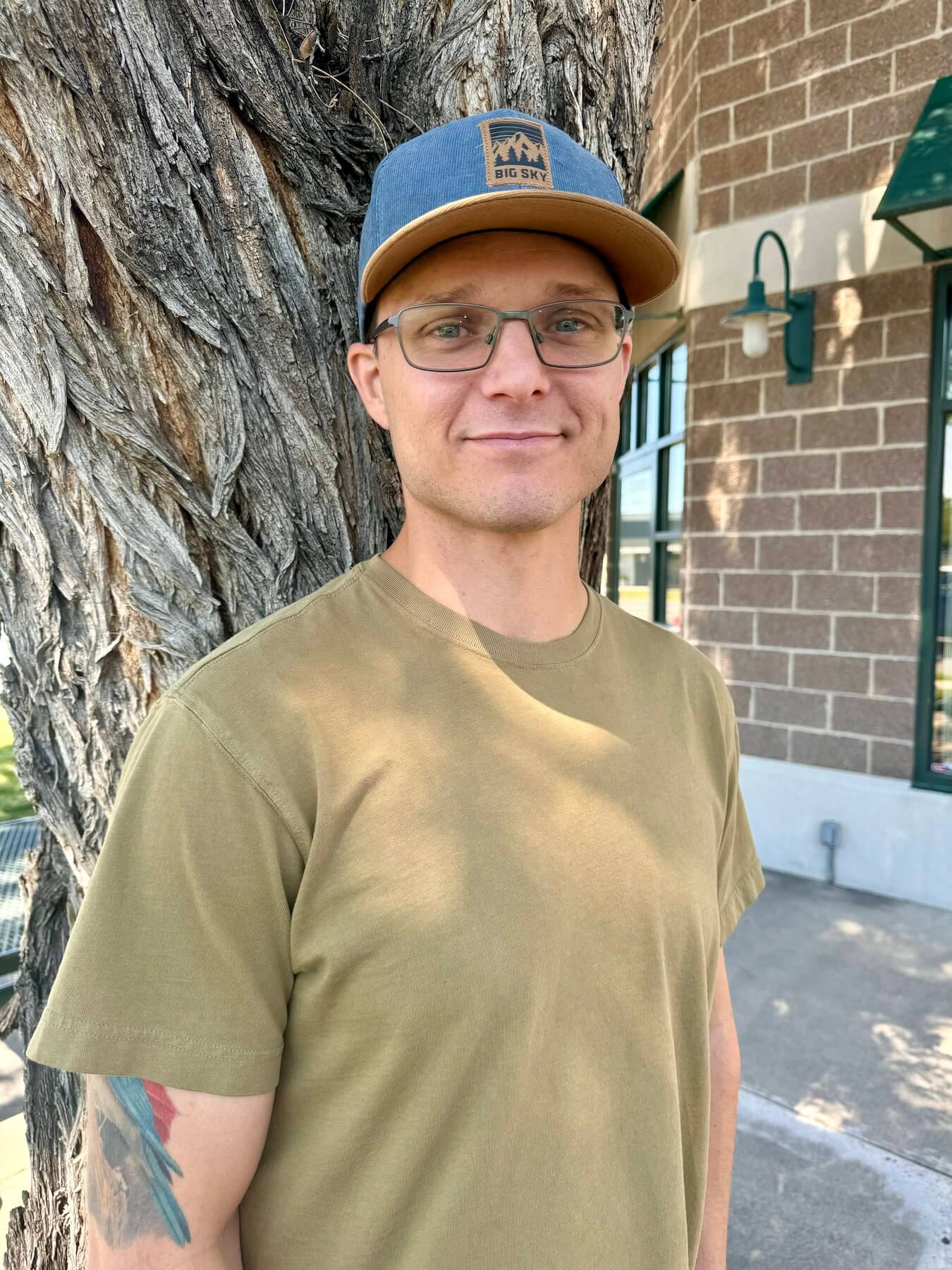 A man wearing glasses and a hat stands in front of a large tree, showcasing a serene outdoor setting.